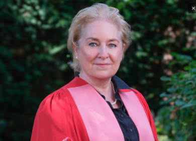 Woman in a red academic gown with a pink stole posing outdoors, greenery in the background.