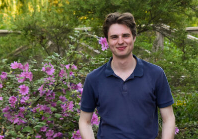 Young man in a navy polo and white pants stands with hands in pockets in a garden with pink flowers.