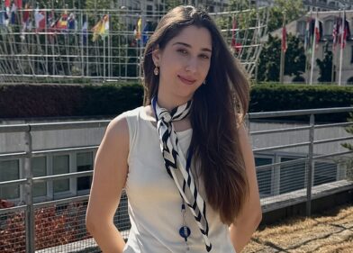 Woman with long dark hair in a white sleeveless top and striped scarf posing outdoors, with a large metal geodesic dome and flags behind her.